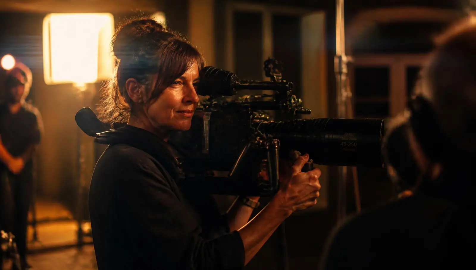 A camera operator filming on a warmly lit production set with crew in the background
